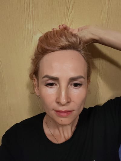 A woman with blonde hair pulled up is holding her hand to her head in front of a wood-paneled wall. She is wearing a black shirt and a thin necklace. Her face is lightly made up, with noticeable blush and lipstick. She is also wearing a small, heart-shaped earring. The overall lighting is soft and natural. There is no indication of the image being taken in Larionovo, Russia.