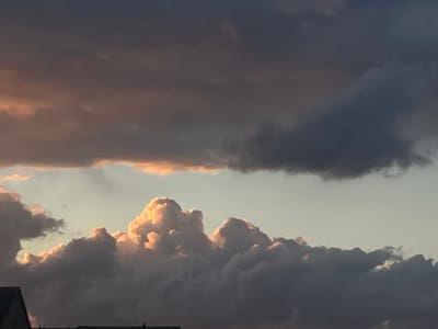 The image captures a dynamic sky over Volodarka, Ukraine, with dramatic clouds illuminated by the setting or rising sun. The clouds are layered, with darker, heavier formations in the foreground and middle ground, casting shadows and creating a sense of depth. Towards the top of the image, lighter, more wispy clouds are tinged with warm, orange and pink hues, suggesting the presence of sunlight breaking through or reflecting off their surfaces. The sky transitions from a pale, almost clear horizon at the bottom center to darker, more ominous tones above. Silhouetted against the sky are the rooftops of buildings in the lower portion of the frame, indicating an urban or semi-urban setting. The overall impression is one of natural beauty and the transient character of atmospheric phenomena.