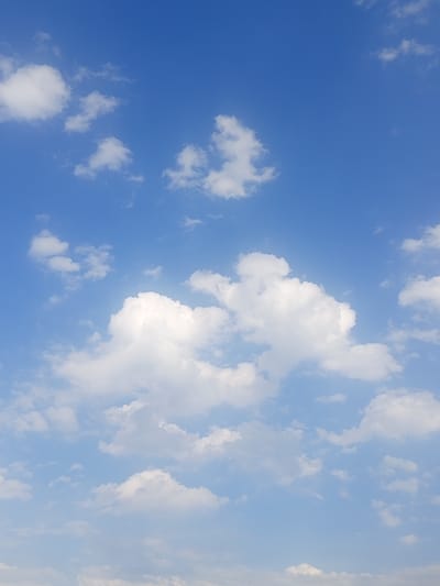 The image depicts a clear blue sky with scattered white cumulus clouds. The clouds vary in size and density, with some appearing wispy and thin, while others are more voluminous and fluffy. The lighting suggests it is daytime, with sunlight illuminating the clouds from above, creating highlights and shadows. The overall scene is serene and atmospheric, characterized by the vast expanse of the sky and the gentle presence of the clouds. The image appears to have been taken from an open area with an unobstructed view of the sky, consistent with a location in Jardy-Su, Kyrgyzstan, which is known for its natural landscapes and clear skies. There are no people, subjects, objects, actions, or interactions visible in the image, other than the natural elements of the sky and clouds.