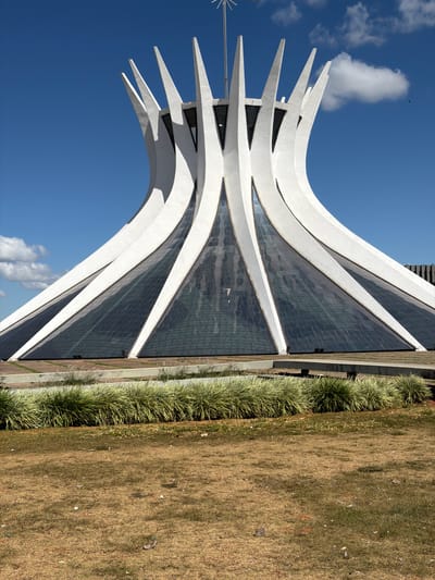 The image captures the Metropolitan Cathedral of Brasília, a hyperboloid structure designed by Oscar Niemeyer. The building features sixteen concrete columns that converge to form a crown-like shape. These columns are painted white and contrasted against the deep blue sky with scattered clouds. The glass panels between the columns create a reflective surface. The Cathedral is surrounded by well-manicured landscaping. The grass is a dry, beige color. A strip of green, bushy plants lines the side of the building. The orientation of the image is rotated.