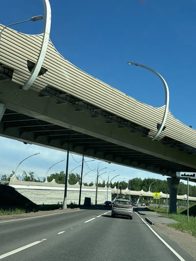 The image displays a forward-facing view from a vehicle on a multi-lane asphalt road in Beloostrov, Russia, under an elevated highway overpass on a clear, sunny day. The dominant structure is the overpass, which features a light-colored, horizontally corrugated or ribbed paneling on its exterior, and multiple decorative, upward-curving white architectural arches with integrated streetlights along its top edge. The underside of the overpass consists of grey concrete support beams and infrastructure.

Below the main overpass, to the right of the road, a secondary elevated structure with a wavy top edge and a horizontally patterned wall is visible. Behind this structure, a line of green trees extends into the background.

On the asphalt road, a grey SUV is positioned in the right lane, moving away from the camera. The vehicle's license plate is obscured. Additional vehicles are visible further down the road in both lanes. The road itself features white dashed lane markings and a solid white line defining the right shoulder, which abuts a concrete curb and a grassy verge.

To the right of the road, a tall, dark pole supports a rectangular white sign with a black silhouette depicting a cityscape, indicative of a city boundary. Further down the road, a yellow triangular sign with a black border and a pedestrian crossing symbol is present. Multiple streetlights are observed, both integrated into the overpass architecture and as standalone poles along the roadway.
