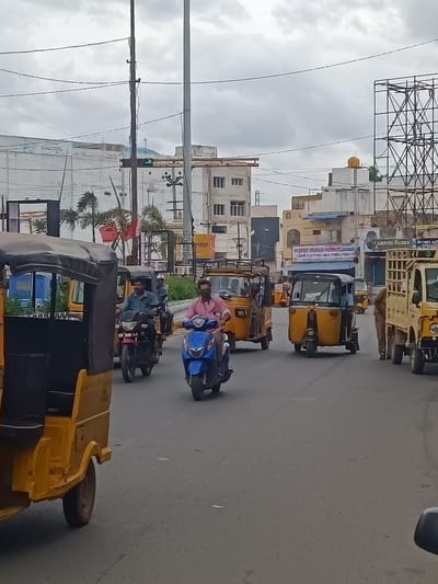 This is a vertical, eye-level, full shot of a street scene in Kurnool, India, under a cloudy sky. The image depicts moderate traffic, primarily consisting of auto-rickshaws and motorcycles. In the foreground, the left side of a yellow auto-rickshaw is visible, taking up a significant portion of the frame. It has a black canopy and a dark interior.
To the right of the auto-rickshaw, several vehicles are in motion. A man in a blue shirt is riding a red motorcycle, heading away from the viewer. Behind him, another person on a motorcycle is partially visible. Further right, a blue scooter is being ridden by a person wearing a pink shirt, also moving away from the viewer. In the background, more auto-rickshaws and a yellow truck with a cargo bed are present. The buildings lining the street are of varying architectural styles, some modern and some older, with signs visible.
One sign prominently reads "VIJAYA DURGA AGENCIES." Another sign, partially obscured, says "GRAND AKBAR." The traffic light in the distance shows a green arrow, indicating traffic is allowed to proceed in that direction. The overall atmosphere suggests a typical day with moderate traffic, possibly during the day given the overcast conditions. The scene is characteristic of an urban or semi-urban environment in India.