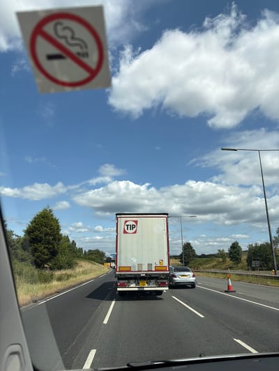 The image depicts a view from a vehicle driving on a highway in Rochdale, United Kingdom. The sky is blue with fluffy white clouds, suggesting a daytime scene with fair weather. The road is asphalt with white lane markings. In the foreground, a square sign with a red circle and a diagonal line over a cigarette symbol, indicating "no smoking," is visible on the left side of the frame. Further down the road, a large white semi-trailer truck with "TIP" logo on its side is traveling ahead. A silver car is also seen in front of the truck. The truck has license plate visible which reads "PJ18 YXM". The truck also has some yellow warning stickers. The road is bordered by a grassy verge and trees on the right. A traffic cone is positioned on the roadside. The perspective suggests the photo was taken looking forward from inside the passenger compartment of a vehicle.