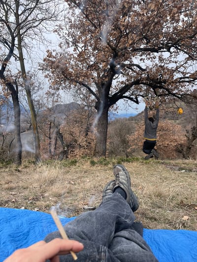 A person wearing jeans and hiking shoes is relaxing on a blue blanket outside with a lit cigarette. In the background, another person hangs from a tree branch with an object in hand. The landscape includes bare trees and hills. The sky is overcast. Smoke from the cigarette drifts into the scene.