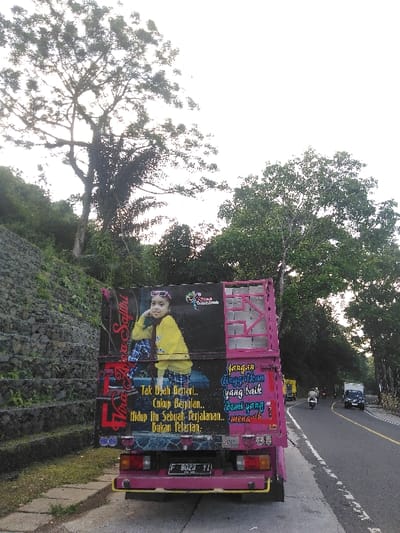 A pink truck is parked on the paved shoulder of a road in Cugenang, Indonesia. The truck's rear features a large graphic of a young girl in a yellow top and pink sunglasses, accompanied by white Indonesian text that translates to "No need to run.. Just walk.. Life is a journey.. Not a sprint." The license plate "F 8023 TI" is visible. To the left of the truck, a vegetated slope with a tiered stone retaining wall ascends, topped by large trees. To the right, the two-lane asphalt road curves slightly, with additional trees lining the roadside. A white motorcycle and two other trucks are visible on the road in the distance, traveling in the same direction as the parked truck faces. The sky is bright with diffused light.
