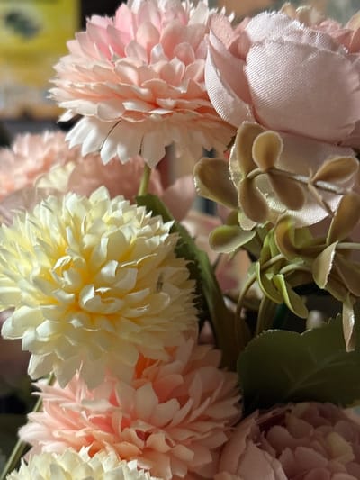 The image displays a close-up view of artificial flowers in soft pastel shades of pink, cream, and light yellow. The flowers have layered petals, creating a full and textured appearance. Some of the flowers are in focus, revealing intricate details of their construction, while others are softly blurred in the background. Interspersed among the flowers are some artificial greenery and small, light brown accents that resemble leaves or buds. The lighting is soft, highlighting the delicate textures of the petals. The setting appears to be indoors, possibly a decorative arrangement. There is no indication of a specific location within Novaya Usman, Russia.