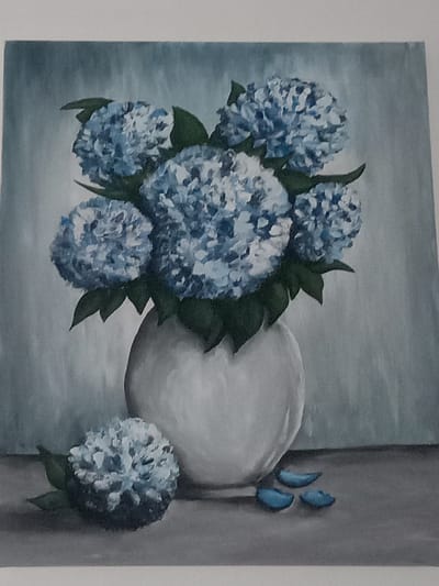 This is an oil painting on canvas, depicting a still life arrangement of blue hydrangeas in a white vase. The vase is round and sits on a flat, grey surface, possibly a table or shelf. Several blooms of hydrangeas are clustered together, with their distinctive flower heads composed of numerous small florets. Some green leaves are visible amongst the flowers. To the left of the vase, a single hydrangea bloom lies on the surface, accompanied by two or three small, detached blue petals. The background is a muted, textured grey-blue, giving the impression of a wall or backdrop. The lighting appears soft and diffused, casting subtle shadows. The painting has a slightly textured, painterly quality. The image does not contain any identifiable landmarks or features that would allow for its location in Kielce, Poland, or any other specific city.