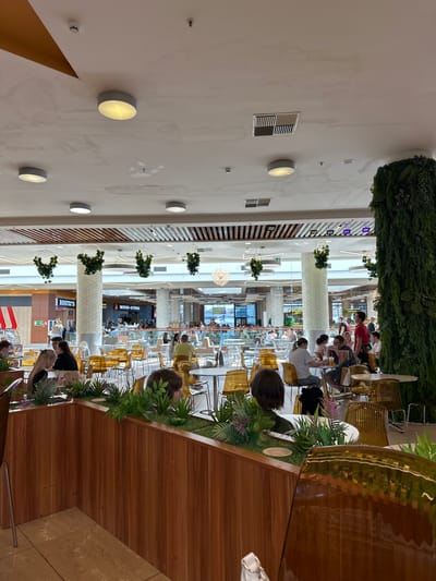 The image depicts an indoor food court in a commercial establishment. The scene is populated with numerous people seated at tables, some engaged in eating or conversing. The environment is characterized by bright overhead lighting, a high ceiling with visible ventilation systems and decorative elements like hanging greenery. Tables and chairs are arranged throughout the space, with some seating areas divided by planters containing artificial foliage. In the background, there are various food stalls or restaurants, indicated by signage. The overall impression is one of a bustling public dining area. The location is identified as Kozulino, Russia.