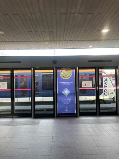 The image depicts a train station platform in Tremblay-en-France, France. A train is visible through glass doors, identified as the CDGVAL, with the car designation "4B" displayed. The platform floor is tiled in gray. Advertisements are affixed to the glass doors, showcasing the "WORLD AIRPORT SKYTRAX AWARDS 2026". Additional text in French and English offers information about Skytrax and Paris Aéroport. The walls are constructed of horizontal stone blocks, illuminated by circular lights.