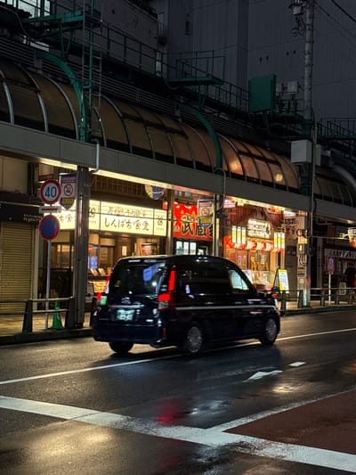 This is a nighttime, street-level, vertical photograph taken in Adachi, Japan. The scene depicts a commercial street with illuminated signs and a dark, wet road reflecting the ambient light. A black vehicle, possibly a car or van, is parked or driving on the right side of the road, its red taillights and side indicators glowing. The left side of the image shows the facade of a building with multiple levels of windows, some of which are lit from within. Various signs in Japanese characters are visible on the building's exterior, with prominent neon lights in yellow and red. One sign reads "しんぱち食堂" (Shinpači Shokudō), indicating a restaurant. Other signs include "北千住" (Kita-Senju), a district in Adachi, and "40" on a speed limit sign. The road surface appears wet, suggesting recent rain or damp conditions, and the white lane markings are clearly visible. The overall atmosphere is one of a bustling urban environment at night, with the lights of businesses and traffic creating a vibrant but somewhat subdued mood due to the darkness and wetness.
