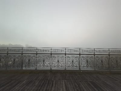 The image displays a waterfront scene along the Schuylkill River in Philadelphia, United States, under conditions of dense fog. In the foreground, a dark, weathered wooden plank deck extends horizontally. Directly beyond the deck, a dark metal railing spans the width of the frame. This railing features an ornate lower section with intricate dark gray scrollwork patterns, topped by a simpler upper section composed of a grid of horizontal and vertical bars. Behind the railing, the surface of the Schuylkill River is visible, appearing calm and reflecting the muted light, its details obscured by the atmospheric conditions. Distant landmasses with indistinct, dark tree lines are faintly discernible on the left, fading into the pervasive, grayish-white fog that severely reduces visibility towards the horizon. The scene is devoid of people, specific subjects, actions, or interactions.