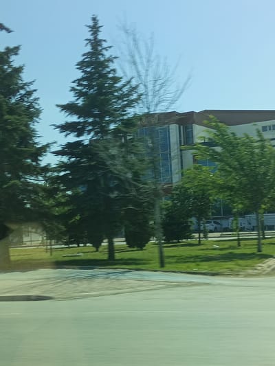 This media file shows a building with a large glass facade and a brown roof, with the word "özel" visible in white text above the glass. The building appears to be a school or office building. It is surrounded by trees and a grassy area, with a road in the foreground. The scene is blurred, suggesting movement, possibly from a vehicle. The sky is clear and blue, indicating a sunny day, likely in the morning or afternoon. The presence of trees and the clear sky suggest a temperate climate. The overall impression is of a modern building in a suburban or semi-urban setting. The motion blur and the focus on the building and surrounding landscape give the image a sense of transience. There are cars visible in the background, parked in front of the building.