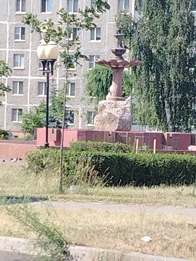 The image captures a sunlit outdoor scene with a multi-story apartment building in the background, characterized by its grid-like pattern of windows. In the foreground, a street lamp with two ornate cream-colored lamps stands to the left. To the right of the lamp, a decorative fountain structure rises, built with rough-hewn stone and featuring tiered basins. This fountain is surrounded by a low, red-brick planter or wall. Dense green foliage, including trees with prominent branches and a trimmed hedge, fills the mid-ground. The ground in the foreground consists of dry, light-colored grass, suggesting a warm climate or season. A paved walkway or road is visible beyond the grass, and a curb or low wall runs along the very bottom edge of the image. A few birds are visible on the ground in front of the red planter. The overall impression is a public space within a residential area. This scene is likely located in Mazyr, Belarus, given the architectural style of the building and the presence of such public amenities.