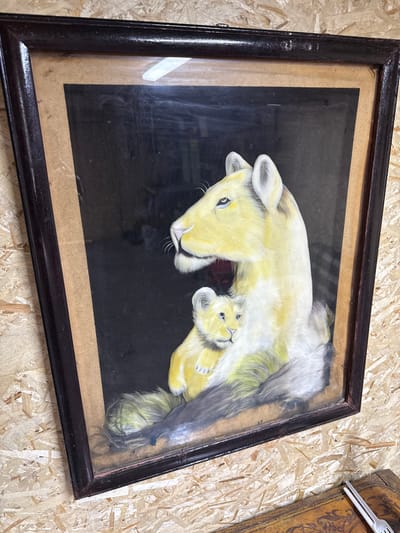 A framed painting of a lioness and cub is displayed on a wooden, particleboard wall. The painting is in landscape orientation, with the lioness in the foreground, her head turned to the left, and her cub nestled against her chest. Both animals are rendered in shades of yellow and white, with dark outlines. The background of the painting is black. The frame is dark brown and appears to be made of wood. The wall behind the painting is made of light-colored particleboard. The image does not provide any information about the city of Айет, Kazakhstan.