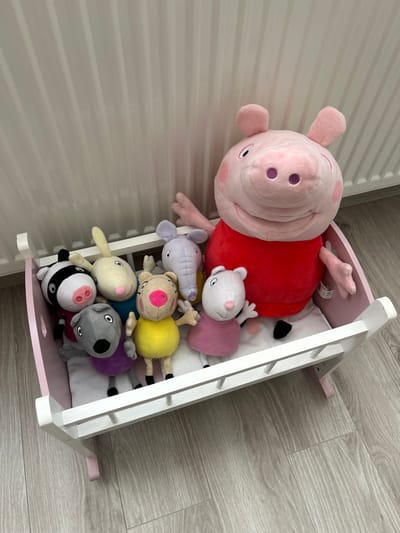 The scene depicts a collection of plush toys arranged inside a toy cradle within an indoor residential setting in Odorheiu Secuiesc, Romania.
A large, pink Peppa Pig plush toy, wearing a red shirt, is positioned at the rear of a white wooden toy cradle, which features light pink side panels and a slatted front rail. Seven smaller plush toys are situated in front of the large Peppa Pig. These smaller toys include a grey elephant, a white rabbit, a grey dog, a beige pig/bear character, a black and white badger/skunk, and a small white mouse/pig. All plush toys appear to be in good condition.
The cradle contains a white padded liner or mattress. The background consists of a plain white wall with vertical ridging, possibly a radiator or a panelled surface. The floor is covered with a light-colored wood-effect laminate or vinyl, displaying a grain pattern in shades of grey and beige. No human subjects are visible.