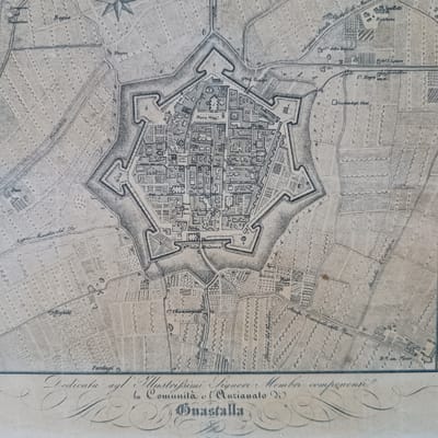 The image is an old map depicting the city of Guastalla, Italy. The city is shown as a star-shaped fortification surrounded by fields, canals, and roads. Within the city walls, a structured layout of streets and buildings is visible, including a "Piazza Maggi" (main square) and "Piazza della Posta". Outside the city walls, various buildings and landmarks are labeled, such as "C. Negri", "Spalletti", "Biolcheria", and "Cimitero degli Ehrei" (Cemetery of the Jews). The text at the bottom indicates the map is dedicated to the "Illustrissimi Signori Membri" (Illustrious Members) of the "Comunità e l'Anzianato di Guastalla" (Community and the Elders of Guastalla). Mezzana Sant'Antonio is not specifically indicated on the map.