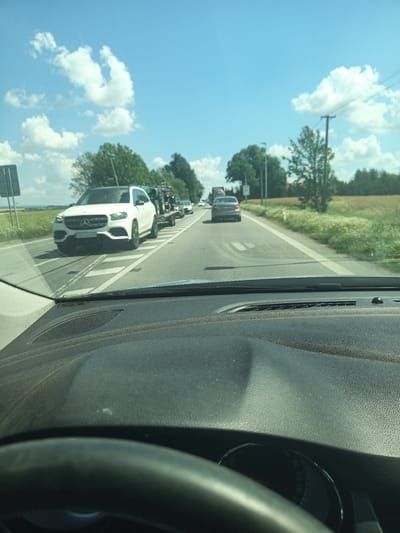 A first-person perspective shot from inside a vehicle shows a white Mercedes-Benz GLS SUV towing a trailer with a vehicle on it. The SUV is driving on a two-lane road in a rural area. The road is asphalt with white lane markings. On the right side of the road, there is a field with tall grass and trees in the distance. On the left side of the road, there are more trees and a blue sky with white clouds. The dashboard of the vehicle is visible in the foreground. The scene appears to be on a sunny day. The license plate of the SUV is visible and reads "ASU 43CP".