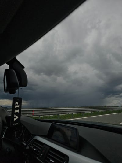 The view from inside a car shows a cloudy sky and a highway. The car appears to be moving on the highway. A rearview mirror and a "VIP" tag are visible within the car. It's impossible to confirm the location as Manolsko Konare, Bulgaria, based solely on the image. There are no people visible.