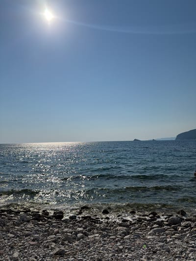The image depicts a sunlit Adriatic Sea coastline in Golubovići, Montenegro. The foreground is a pebble beach with smooth, grey and white stones of various sizes. The water is a deep blue, with the sun's bright reflection creating a shimmering pathway across its surface. Gentle waves lap at the shore. In the distance, hazy mountains form the horizon, with a small island visible to the left. The sky is clear and bright blue, with the sun positioned in the upper left quadrant, casting a strong glare. A portion of a person's head and shoulder are visible in the lower right corner of the frame, suggesting they are in the water.