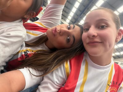 Three females are pictured in an indoor setting. The girl on the left has pale skin and light brown hair, while the girl in the center has darker skin and brown hair. The third girl is partially visible at the bottom right corner of the image. All three are wearing white t-shirts with red and yellow stripes. The background appears to be a building's ceiling with metal beams and fluorescent lighting. It's impossible to determine the exact location within Tlaxcalancingo, Mexico from the given information.