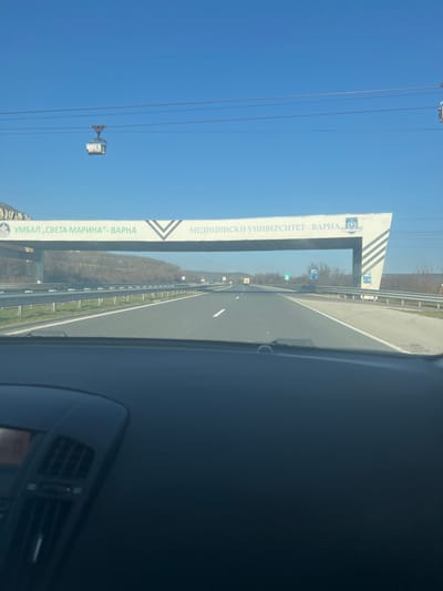 This image depicts a multi-lane asphalt highway on a clear day, viewed from the interior of a vehicle, evidenced by the dark dashboard occupying the lower third of the frame. The scene is located on a major highway in Bulgaria, specifically the A2 Hemus highway, within Varna Province. This position is on the approach to Varna, approximately in the area of the village of Kipra, which is also located in Varna Province.
The dominant feature is a white, angular pedestrian overpass or banner structure spanning the entire width of the highway. On the left side, the structure displays the text "УМБАЛ „СВЕТА МАРИНА“- ВАРНА" (UMBAL "SVETA MARINA" - VARNA) along with a green circular logo. On the right side, it reads "МЕДИЦИНСКИ УНИВЕРСИТЕТ - ВАРНА" (MEDICAL UNIVERSITY - VARNA) next to a blue crest-like logo.
Above the overhead structure, a cable car system is visible against a bright blue sky. One white gondola, numbered '5', is suspended from overhead cables on the left side of the frame, appearing to move horizontally. Another cable support structure is partially visible on the far right.
The highway has visible white lane markings and is flanked by metal guardrails. In the distance, several other vehicles are discernible on the road, including a light-colored truck. Distant hills or low mountains with bare trees line the horizon on both sides of the highway, indicating a rural or semi-rural environment. Small blue and white road signs are also visible on the right side of the highway beyond the main overhead structure. No specific individuals are identifiable within the frame. The perspective suggests active travel.
