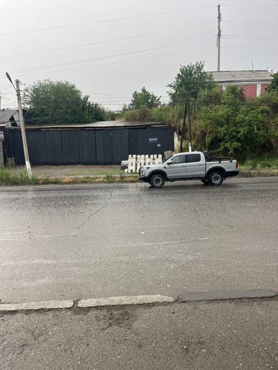 A silver pickup truck is parked on the wet asphalt of a road, reflecting the overcast sky. The truck is positioned on the right side of the frame, facing left. In the background, a dark metal corrugated fence runs horizontally across the image, with a red and white building visible above it. Lush green trees and vegetation line the area behind the fence. Power lines stretch across the sky, and a lamppost stands to the left of the truck. The road is wet and appears to have some cracks and repairs. The scene suggests an outdoor, possibly industrial or commercial area in Medrese, Kyrgyzstan, under cloudy weather conditions.