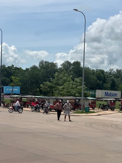 This image captures a scene in Bavet Quarter, Cambodia, likely a transportation hub or border crossing area. In the foreground, two men stand on a paved surface, one wearing a light-colored hat, light shorts, and a long-sleeved shirt, the other in a plaid shirt and dark pants. To their left, several motorcycles are parked and in motion. One man on a motorcycle wears a white head covering and a light-colored shirt. Behind them, a row of tuk-tuks, small three-wheeled vehicles, are lined up, some with passengers. In the background, a dense line of green trees forms a backdrop. To the left, a blue shipping container with "MAERSK" written on it is visible. To the right of that, a white shipping container with "Matson" printed on it. Tall streetlights with modern fixtures are also present. The sky is partly cloudy with large white cumulus clouds. The ground shows tire marks from vehicles.