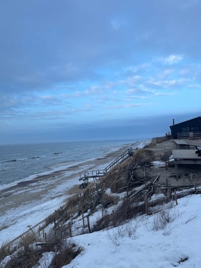 The image captures a winter coastal scene in Morskoe, Russia. A vast body of water, likely the Baltic Sea, extends across the left half of the frame under a partially cloudy, blue-grey sky. The shoreline consists of a sandy beach, extensively covered with white snow and ice. To the right, a snow-dusted sandy dune or bluff rises, sparsely vegetated with dry, brown grasses and bare shrubs. Multiple wooden posts and horizontal timbers form erosion control structures along the dune face. Two distinct wooden staircases with railings descend the dune towards the beach level. On the upper right, situated atop the dune, stands a dark-colored building with a sloped roof, multiple windows, and a chimney stack. Adjacent to the building is an elevated wooden deck or platform. The immediate foreground is covered in snow with small, dark shrubbery visible. No individuals or animals are present in the scene.