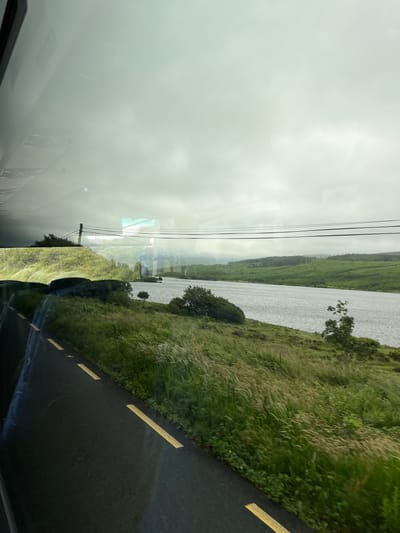 The image shows a view from inside a vehicle, looking out at a cloudy sky over a body of water and a lush green landscape. A dark asphalt road with yellow dashed lines is visible in the foreground, suggesting the perspective is from a car or bus traveling along a road. The landscape includes rolling hills covered in grass and trees, with a wide river or lake in the middle ground. In the distance, more hills and trees are visible, shrouded in a hazy atmosphere. Overhead, power lines stretch across the frame, supported by a distant pole. The overall impression is of a scenic, rural location in Ireland, likely the Gweedore area, known for its natural beauty and coastal landscapes. There are no people or specific structures evident that would pinpoint an exact location within Gweedore, but the environment is consistent with the region.