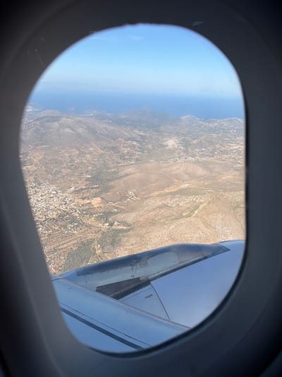 An aerial view captures a landscape from the window of an airplane, revealing a sunlit panorama of Yagel, Israel. The image is significantly tilted, as if the aircraft is banking, with the horizon appearing vertically on the left side of the frame.
Through the elliptical window, a vast expanse of arid, rocky terrain dominates the foreground and midground, characterized by sparse scrubland and scattered patches of greener vegetation, likely olive groves or other cultivation. Interspersed throughout this landscape are numerous small buildings and clusters of settlements, connected by visible roads and tracks, indicative of human activity in the region.
To the far left, a distinct horizon line separates the land from a calm, blue body of water, likely the Mediterranean Sea, stretching into a clear, light blue sky above. The scene is illuminated by bright daylight, suggesting a clear and sunny day, possibly midday or early afternoon, with no clouds visible.
On the right side of the frame, a significant portion of the airplane's wing and engine cowling is visible. The metallic surfaces of the engine reflect the bright sky, with various panels and structural elements clearly defined. Notable text visible on the engine cowling reads "JANICE SIMON." Further back on the wing, near the engine pylon, a small white rectangular label with "10 17" is discernible. No people are visible in the image.