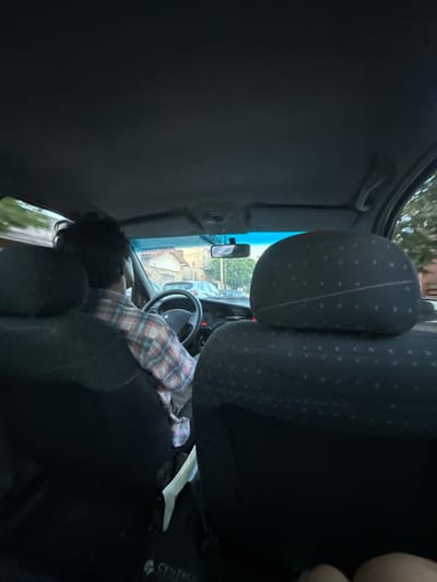 The image captures the interior of a vehicle from a rear passenger's vantage point, looking forward onto a street in Villa Yapeyú, Argentina. A male driver, with dark, curly hair, is positioned in the driver's seat, wearing a plaid shirt patterned with red, white, and blue checks. His hands are on the black steering wheel, which features a central emblem. The vehicle's interior presents a dark headliner and dark fabric seats. The headrest of the front passenger seat is visible on the right. Between the two front seats, a vertical white object is discernible. In the lower right corner, the knees of a second rear passenger are visible. Through the windshield, a street scene unfolds under what appears to be late afternoon or early evening light. On the left side of the street, residential buildings with light-colored walls and visible tiled roofs are present. Parked vehicles, including a silver sedan, line the street. Green foliage from trees is visible along the roadside and in the background, alongside utility poles and overhead wires.