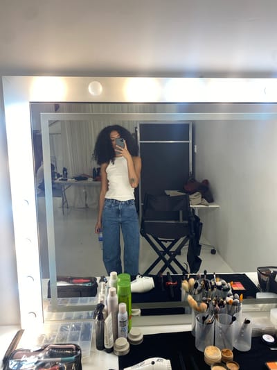 The image shows a person reflected in a makeup mirror with lights around the frame. The person is holding a smartphone and taking a picture of themselves. The person is wearing a white tank top and blue jeans. The mirror is surrounded by makeup brushes, bottles, and other beauty products. In the reflection of the mirror, there is a makeup chair and a backdrop. The location is likely a makeup studio or salon. There is no indication of the city of Porto Alegre, Brazil.