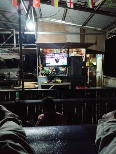 The image depicts an outdoor viewing area at night, likely in Mesjidpirak, Indonesia, with several individuals observing a television screen. The setting appears to be a casual establishment, possibly a warung or small cafe, with a corrugated metal roof supported by metal poles. Flags of various nations are strung across the roofline, suggesting a sporting event is being broadcast.
In the foreground, the legs of at least two individuals are visible, indicating they are seated or reclining on a dark, textured surface. Behind them, two other individuals are seen from the back, one in a striped shirt and another facing away from the camera. One of these individuals appears to be holding a smartphone.
The central focus of the scene is a flat-screen television displaying a close-up of a dark-skinned man with a beard, likely a basketball player given the context of athletic imagery. Below the television, a counter or shelf displays numerous bottles, possibly beverages, in various colors including green and red. Behind the counter, shelves are stocked with goods, and a blue door is slightly ajar. The overall lighting is dim, with the television and a light bulb under the roof providing the primary illumination. The environment is rustic, with exposed wooden structures and a dark, wooden fence-like barrier in the middle ground. A sign with Indonesian text, including "BANJIR ACEH UTARA", is partially visible on the left side, indicating a connection to a flood in North Aceh.