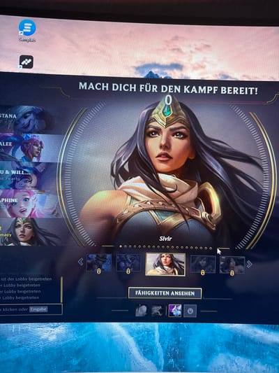 A computer screen, located in Vellmar, Germany, displays a video game interface (likely League of Legends) and a portion of a desktop background. In the upper-left corner of the screen, two desktop icons are visible: the top one is a blue circular icon with three horizontal white lines and two white arrows, labeled "Samplab." Below it is a black square icon with a white, abstract, arrow-like symbol. The desktop wallpaper behind these icons depicts a serene landscape with soft pink and orange clouds against a light blue sky, above distant, hazy blue-grey mountains. The primary focus is the game interface, featuring a large, central portrait of the character "Sivir." She has long, dark hair, dark eyes, and a determined expression. She wears a gold and teal headpiece adorned with a prominent green emerald-like gem, and matching armor that reveals her shoulders. Above her headpiece, a green "0" is displayed. Sivir's portrait is enclosed within a gold circular frame with numerous small vertical white tick marks. Below her portrait, her name "Sivir" is clearly visible, followed by a horizontal row of small circular indicators, some filled, some empty, likely representing different character skins. A white mouse cursor, shaped like an arrow, points towards the right-most of these circles. Above Sivir's portrait, a dark banner carries the German text: "MACH DICH FÜR DEN KAMPF BEREIT!" (English: "GET READY FOR BATTLE!"). A slight visual artifact, resembling a faint yellow-green line, is present above the 'CH' in 'MACH'. To the left of Sivir's portrait, a vertical list of smaller character portraits is partially visible, indicating a character selection menu. Visible names include truncated sections like "STANA," "ALEE," "U & WILL...", "APHINE," and a smaller Sivir portrait at the bottom of the list. Below the main Sivir portrait, a horizontal row displays smaller icons representing character skins; Sivir's primary skin is highlighted, flanked by darkened, locked skins marked with a gold '8'. Below this row, a button reads "FÄHIGKEITEN ANSEHEN" (English: "VIEW ABILITIES"). Further down, a row of four small square ability icons are present: the first two are grey and indistinct, the third is a stylized purple and blue butterfly-like spell effect, and the fourth is a grey square containing a white clock icon. On the bottom-left of the game interface, a chat or log window displays multiple lines of German text, "ist der Lobby beigetreten" (English: "has joined the lobby"), with a vertical