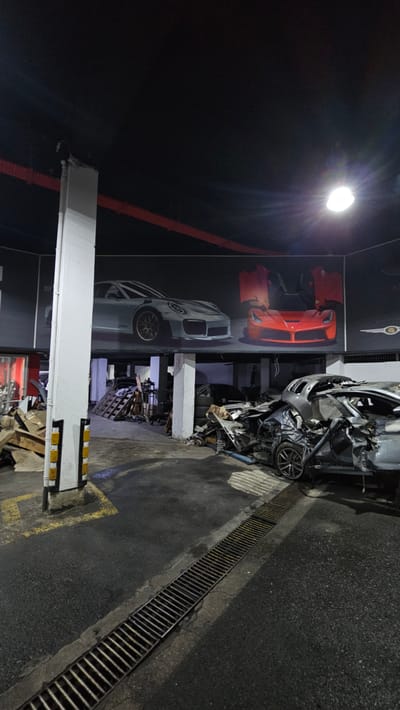 The image depicts a dimly lit, industrial-style garage or workshop interior, potentially located in Ataşehir, Turkey. The scene is characterized by a stark contrast between a large mural of pristine sports cars adorning the back wall and the foreground which is filled with debris and heavily damaged vehicles.

The mural features two prominent sports cars: a silver Porsche on the left and a vibrant orange Ferrari on the right, both with their doors open, creating an illusion of luxury and speed. This mural is mounted high on the wall, above the working area of the garage.

In the foreground, the floor is a rough, dark asphalt surface with a prominent metal grate drain running horizontally across the lower half of the image. Scattered throughout the space are various automotive parts and what appear to be wrecked car bodies. Most notably, a severely damaged silver car, possibly a sports car, is situated in the middle-right of the frame. Its front end is crushed, and the body panels are heavily deformed, indicating a significant collision. Other debris, including wooden pallets, tires, and metal scraps, are piled around this damaged vehicle and in other areas of the garage.

A tall, white structural pillar with yellow and black protective bollards is visible on the left side of the image, partially obscuring the view of some of the debris. The ceiling is dark and appears to have exposed pipes or conduits, with a bright industrial light fixture illuminating the upper right portion of the scene. The overall impression is one of a repair shop that handles significant vehicle damage, with the juxtaposition of the idealized mural and the harsh reality of wrecked cars. There are no people visible in the image.