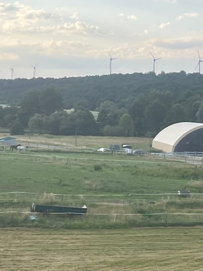 The image depicts a rural landscape with several wind turbines visible on a distant hill under a cloudy sky. In the foreground, there is a fenced pasture with two horses, one white and one dark. Further back, a large, arched building with a light-colored roof is visible. A dark-colored trough or water feeder is present in the middle ground, lying on its side within a fenced area. The scene suggests a farm or equestrian facility located in a rural setting, possibly near Momberg, Germany, given the wind turbines common in German landscapes.
