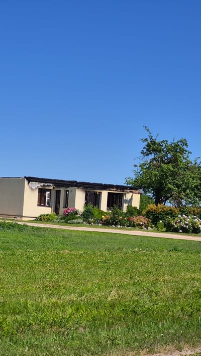 The image depicts a one-story, cream-colored building situated in Astašova, Latvia. The building appears to have sustained significant fire damage, with portions of its roof charred and missing, exposing the dark structure beneath. The windows are dark, and some appear to be damaged. In front of the building, a variety of flowers and shrubs are planted, with pink and white blooms visible. To the right of the building, a large, mature tree with green foliage stands tall. The foreground is dominated by a lush green lawn, with a dirt path running horizontally across the middle ground. The sky above is a clear, bright blue. There are no people or animals visible in the image.