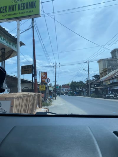 The image captures a street view in Kotapantonlabu, Indonesia. On the left side of the road, a sign reads "CEK MAT KERAMIK PANTON LABU - ACEH UTARA," indicating a ceramic material shop. Adjacent to it, another sign says "POLSEK" with an arrow pointing left and "50 M," suggesting a police station is nearby. Several stacks of ceramic tiles are visible in front of the shop. The street itself appears to be a two-lane road with moderate traffic, including motorcycles and a green bus in the distance. Buildings line the right side of the road, featuring a mix of commercial establishments with corrugated metal roofs and more modern concrete structures. Overhead, a network of power lines crisscrosses the sky. The sky is partly cloudy, suggesting it is daytime. The perspective is from inside a vehicle, looking forward through the windshield.