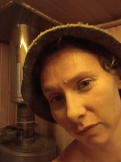 The image shows a close-up of a woman wearing a dark gray sauna hat. She is in a sauna, indicated by the wooden walls and a visible stainless steel sauna stove. The lighting is warm and yellow, characteristic of a sauna. The woman is looking towards the camera with a neutral expression. Her skin appears flushed, possibly from the heat of the sauna. The background shows the interior of a sauna, with the stove and wooden paneling prominently featured. The exact location in Verkhneye Dubrovo, Russia cannot be determined from the image alone.