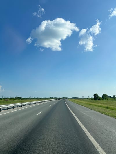 The image depicts a view from a vehicle traveling on a multi-lane highway in Verkh-Tula, Russia. The road is asphalt, with clear white lane markings. A metal guardrail runs along the left side of the highway, separating it from a grassy verge. Beyond the verge, there are fields, trees, and utility poles. The sky is a clear blue with scattered white clouds. In the distance, a few vehicles can be seen on the highway. The overall impression is of a bright, sunny day on a rural highway.