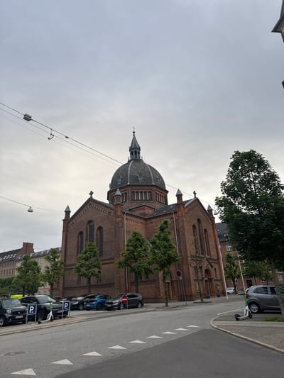 The image shows the Grundtvig's Church in Frederiksberg, Denmark, under an overcast sky. The church is a large, red brick building with a prominent dome and a spire. In front of the church, there is a street with parked cars and two electric scooters. One scooter is green and white and is parked near the curb, while another is partially visible behind a parked car. The street is marked with white arrows indicating a pedestrian crossing. Trees line the street and the front of the church. The environment appears to be an urban setting.