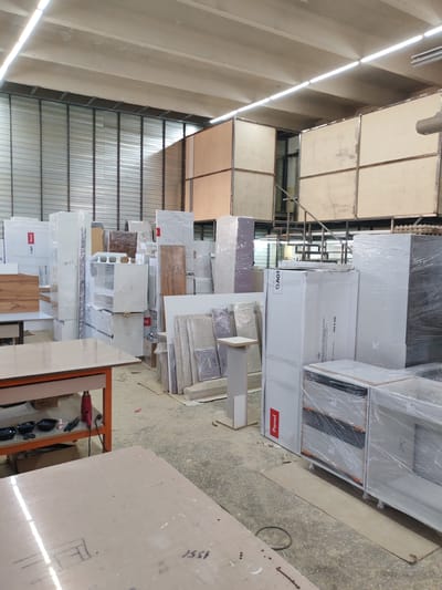 The image shows the interior of a warehouse or storage facility in Bratsk, Russia. The space is filled with stacks of furniture components, likely for kitchens or wardrobes, wrapped in plastic. Tall, white cabinet units are prominent, some with labels indicating brand names like "DAGT" and "Panel". There are also flat panels, possibly cabinet doors or shelving, leaning against each other.
In the foreground, a workbench with orange metal legs is visible, holding various tools including a red heat gun, a glue bottle, and bowls containing small dark objects. A light-colored table surface is in the lower right corner of the image, with some handwritten markings. The floor is concrete, with some scattered debris and cardboard pieces. In the background, metal shelving units and large wooden panels are stacked high, reaching towards the ceiling. The overall environment suggests an active manufacturing or distribution center for furniture.