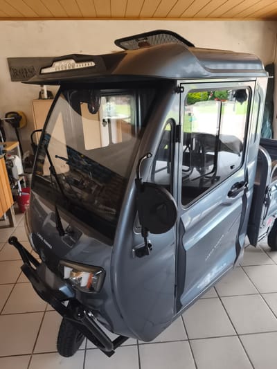 This media file shows a close-up, front-right view of a gray, three-wheeled electric cargo vehicle, specifically a "CARGO RUNNER". The vehicle appears to be parked indoors, possibly in a garage or workshop, with a tiled floor and a wooden-slatted ceiling visible. The setting suggests it's a private space.
The vehicle itself is the primary subject. It features a closed cabin with two doors, large windshields, and side windows that reflect the outdoor environment. The exterior is a glossy gray. Notable details include the front bumper, headlights, windshield wipers, and side mirrors. The text "CARGO RUNNER" is visible on the side of the vehicle, along with a website address: "www.e-mobile-hilusch.de". There is also a prominent light fixture above the windshield, featuring several illuminated circles and the text "IN PENG". A small fan is mounted inside the cabin, to the left of the driver's side.
There are no people visible in the image, and the activity taking place is simply the vehicle being stationary. The lighting suggests it is daytime, but the indoor setting makes it difficult to determine the exact time of day or weather conditions outside. The location context provided is Bad Iburg, Germany, which is relevant to the website address. The overall impression is one of a well-maintained, modern electric vehicle presented in a clear, focused shot.