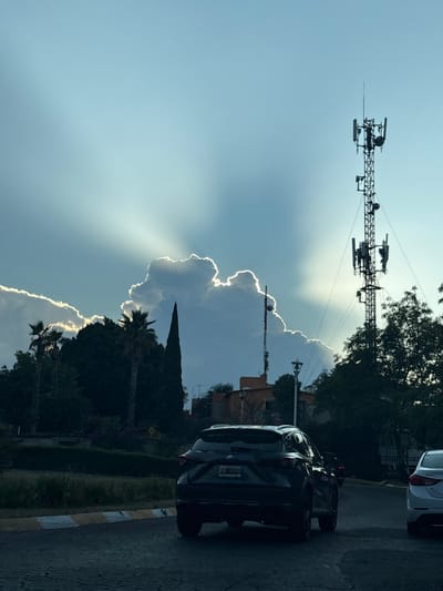 A cell tower rises against a bright sky with sun rays filtering behind a large cloud formation. Trees and a tall, pointed evergreen fill the lower portion. Cars are parked on the right side of the road, which has a yellow and white striped curb.