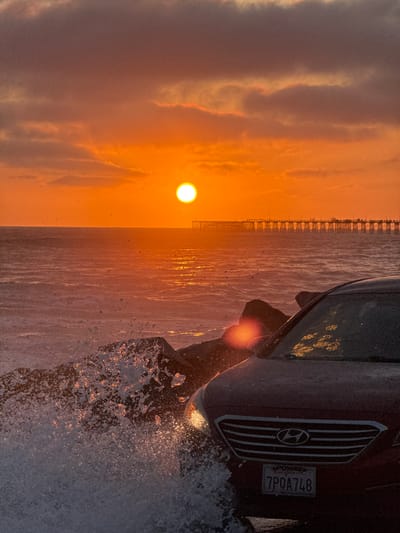 A red Hyundai Sonata is parked near a rocky shoreline during sunset in Oceanside, California. The sun is a bright orb low on the horizon, casting a warm orange glow across the sky and the ocean. Clouds are present in the sky, with some appearing dark and others tinged with orange. A long pier stretches across the ocean in the distance. Waves are crashing against the rocks and splashing onto the front of the car. The car's license plate is visible and reads "7PQA748". There are no people visible in the image.