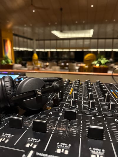 The image shows a close-up of a DJ mixer and a pair of headphones. The mixer is black with many knobs, faders, and buttons. Several faders have numerical markings from 0 to 10, and some have labels like "SEND" and "CUE." Small LED lights are visible above some of the controls. The headphones are also black, with the Sennheiser logo on the earcups.

In the blurred background, there appears to be an indoor setting with wooden paneling on the walls and ceiling. There are also some lights visible in the background, possibly part of the decor or lighting system. A yellow decorative object, possibly a lamp or vase, is visible in the far left background, along with some greenery. The overall lighting suggests an indoor environment, possibly at night or in a dimly lit space. The image is taken at an angle, with the DJ equipment dominating the foreground. There is no indication of people or any specific activity taking place other than the implied use of the DJ equipment. The location context provided ("Unknown, Maldives") is not visually supported by the image content itself.