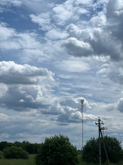 The image depicts a rural landscape under a cloudy sky. A tall, slender telecommunications tower stands prominently in the middle ground, with multiple antennas arrayed at its apex. To its right, a wooden utility pole supports electrical lines and insulators. In the foreground and stretching across the lower portion of the image is a dense line of lush green trees and foliage. The ground appears to be covered in grass, suggesting a natural or agricultural setting. The sky is a mix of blue patches and voluminous, greyish-white clouds, indicating dynamic weather conditions. The scene appears to be located in or near Старое Бобренево, Russia, consistent with its rural characteristics. There are no people or animals visible in the image.