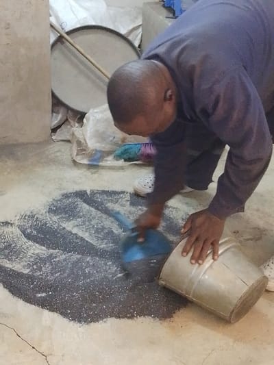 In an indoor setting within Olomoda, Nigeria, a dark-skinned adult male is depicted actively scooping a dark, granular material from a cracked concrete floor. The man, with a close-cropped haircut, wears a dark blue long-sleeved shirt, dark pants, and white slip-on shoes. He is bent forward, using a blue plastic scoop held in his left hand to gather the material. His right hand rests on the rim of an overturned, silver-colored metal bucket, positioned to collect the scooped contents. The dark granular material, intermixed with lighter dust, is spread in an irregular pattern on the floor. In the background, against a light-colored wall or pillar, are white sacks, a large circular flat metal object with a wooden handle leaning against it, and a multi-colored brush on the floor.