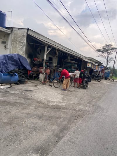 The image depicts a street scene in Kecamatan Margaasih, Indonesia, a small town located in the Bandung Regency. The scene is captured from a perspective that looks down the street, with the camera positioned in front of a roadside shop. The shop, which appears to be a motorcycle repair or tire shop, has a weathered concrete facade and an open front. Stacks of tires can be seen inside the shop, and there are also a few items of machinery, including an air compressor and a workbench. Several people are gathered around the shop. Two men stand in front of the shop, one in a red shirt and the other in a blue shirt. They appear to be talking. A third man, also in a red shirt, is crouched down beside a bicycle with its back wheel removed. He is working on the bike. Two other men stand behind the first two men, watching the man with the bike. They are wearing casual clothing. The street is made of asphalt, and it is largely empty. Several power lines run overhead, and there is a small tree in the distance. The sky is overcast, and the overall atmosphere is subdued.