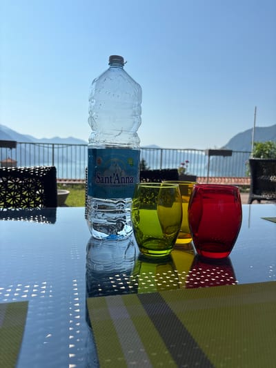 The image shows a still life arrangement on a glass table with a backdrop of a sunny sky and distant mountains. The primary subjects are a plastic bottle of Sant'Anna Acqua Minerale Naturale and three colored glasses. The bottle is clear and filled with water, with condensation visible on its surface. The label on the bottle reads "Sant'Anna," "ACQUA MINERALE NATURALE," and "SORGENTE OSPEDALE 1930m." Two of the glasses are filled with a yellow liquid, and one is filled with a red liquid. These glasses are positioned behind the bottle.

In the background, there is a railing and some greenery, suggesting an outdoor setting, possibly a balcony or patio overlooking a landscape. The sunlight is bright, creating distinct shadows and reflections on the glass table, including a pattern of triangular light shapes cast by the placemat. The time of day appears to be late afternoon or early evening, given the angle of the sunlight and the clear blue sky. There are no people visible in the image. The overall mood is serene and inviting, perhaps suggesting a moment of refreshment outdoors. The scene is set outdoors with a mountainous landscape in the distance, possibly in Zorzino, Italy, as indicated by the context.